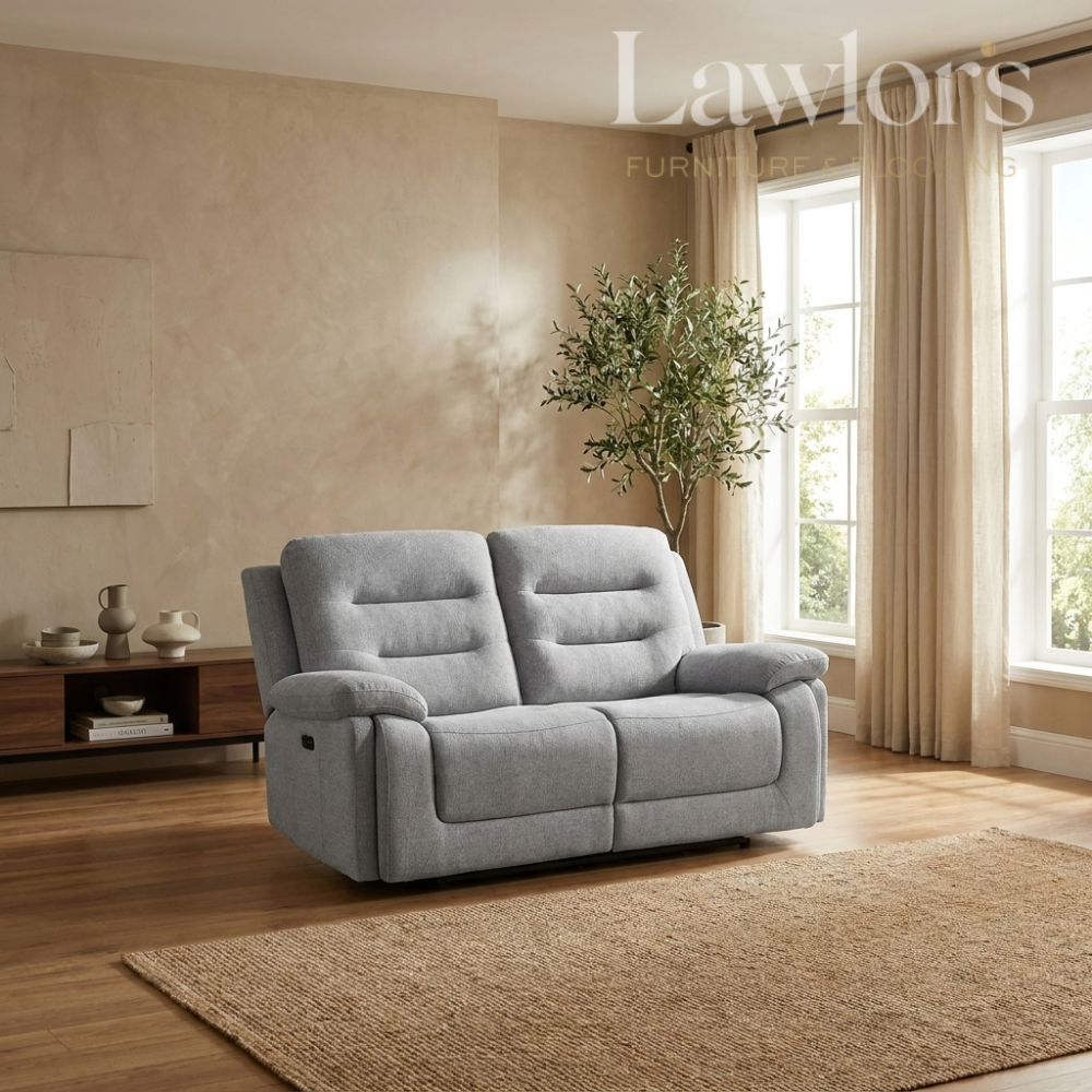 A light gray upholstered reclining loveseat sofa shown in a styled living room with wood floors, beige walls, curtains, a plant, and a rug.