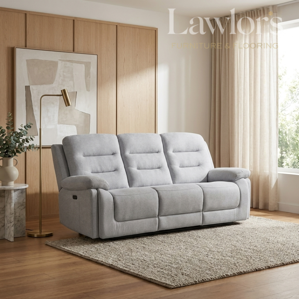 A light gray three-seat reclining sofa in a modern living room setting with wood paneling, a floor lamp, a side table with a vase, and a large window with sheer curtains.