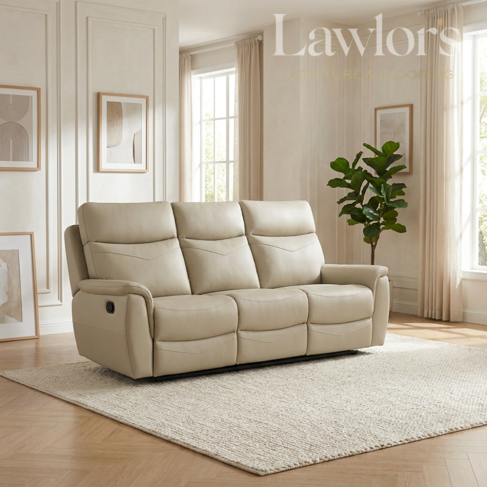 A cream-colored three-seater reclining leather sofa placed on a textured light rug in a bright, modern living room. The room features beige walls, framed abstract art, a potted green plant, and large windows with beige curtains.