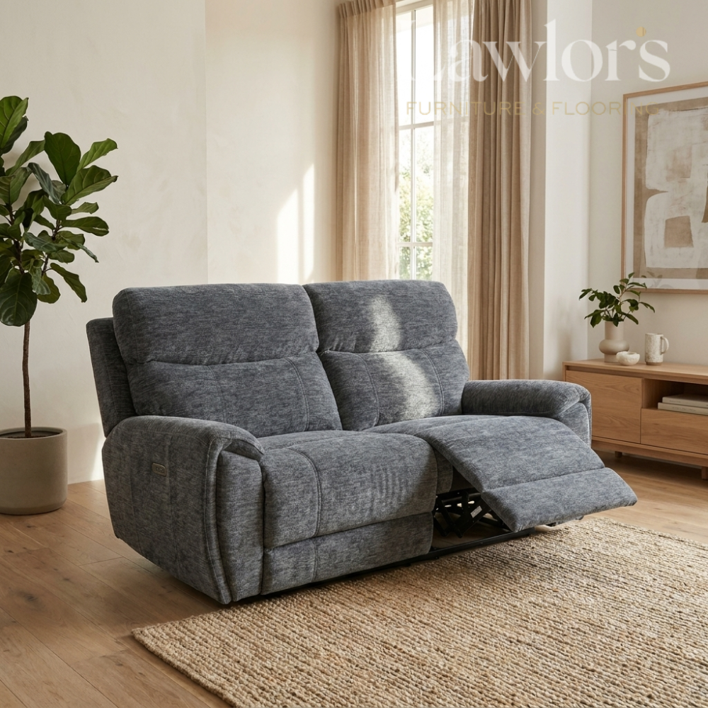 A gray upholstered reclining loveseat in a bright living room, with the front cushion extended. The room has beige walls, a window with sheer curtains, a wooden side table, a plant, and a woven rug.