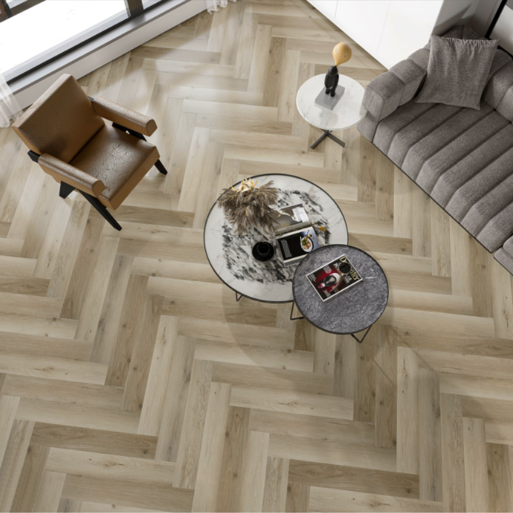 Light oak herringbone wood flooring shown in a modern living room setting with a brown leather armchair, a gray sofa, and two round nesting coffee tables.