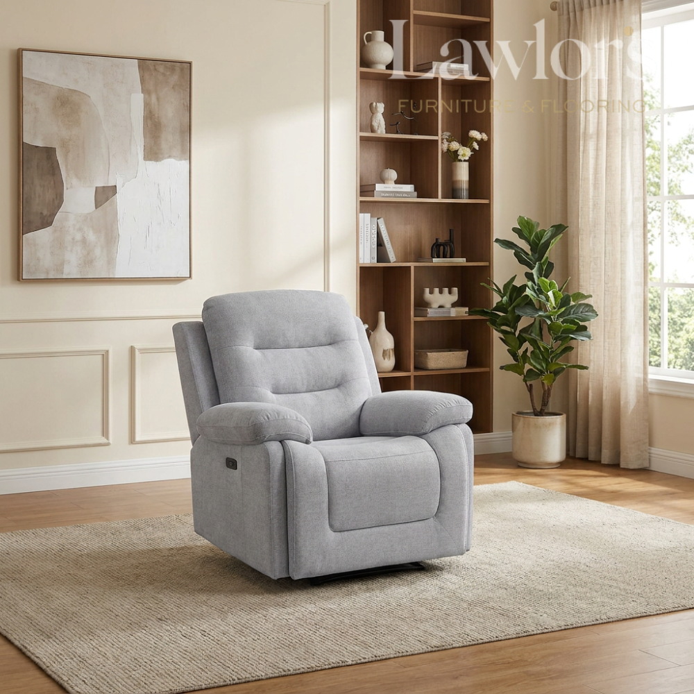 A light gray upholstered recliner chair placed on a beige rug in a bright living room with wood flooring, a bookshelf, wall art, and a potted plant.