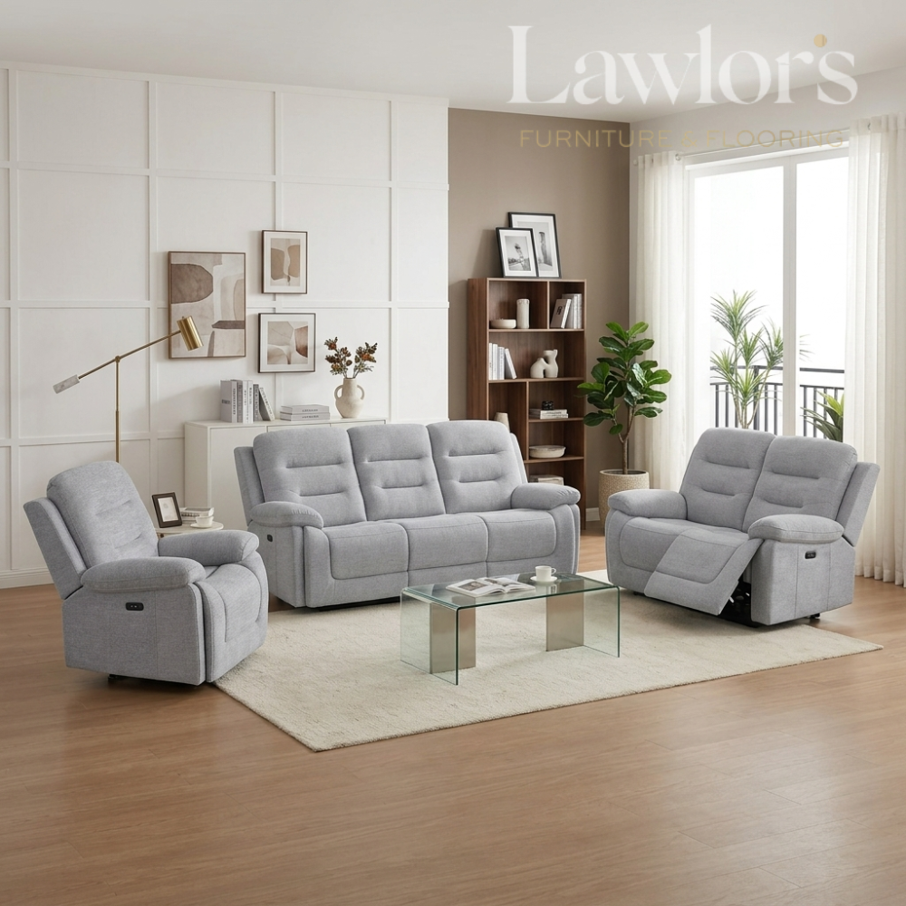 A light gray three-piece reclining living room furniture set including a sofa, two armchairs, and a glass-top coffee table, staged in a modern living room with wood floors, a white rug, and a window.