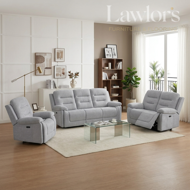 A light gray three-piece reclining living room furniture set including a sofa, two armchairs, and a glass-top coffee table, staged in a modern living room with wood floors, a white rug, and a window.