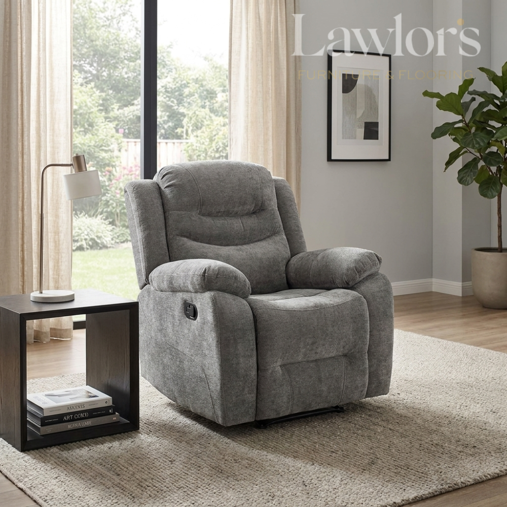 A gray fabric recliner chair in a modern living room setting, positioned next to a black side table with a lamp and books. The room features light gray walls, a beige rug, and a large window with curtains.