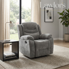 A gray fabric recliner chair in a modern living room setting, positioned next to a black side table with a lamp and books. The room features light gray walls, a beige rug, and a large window with curtains.