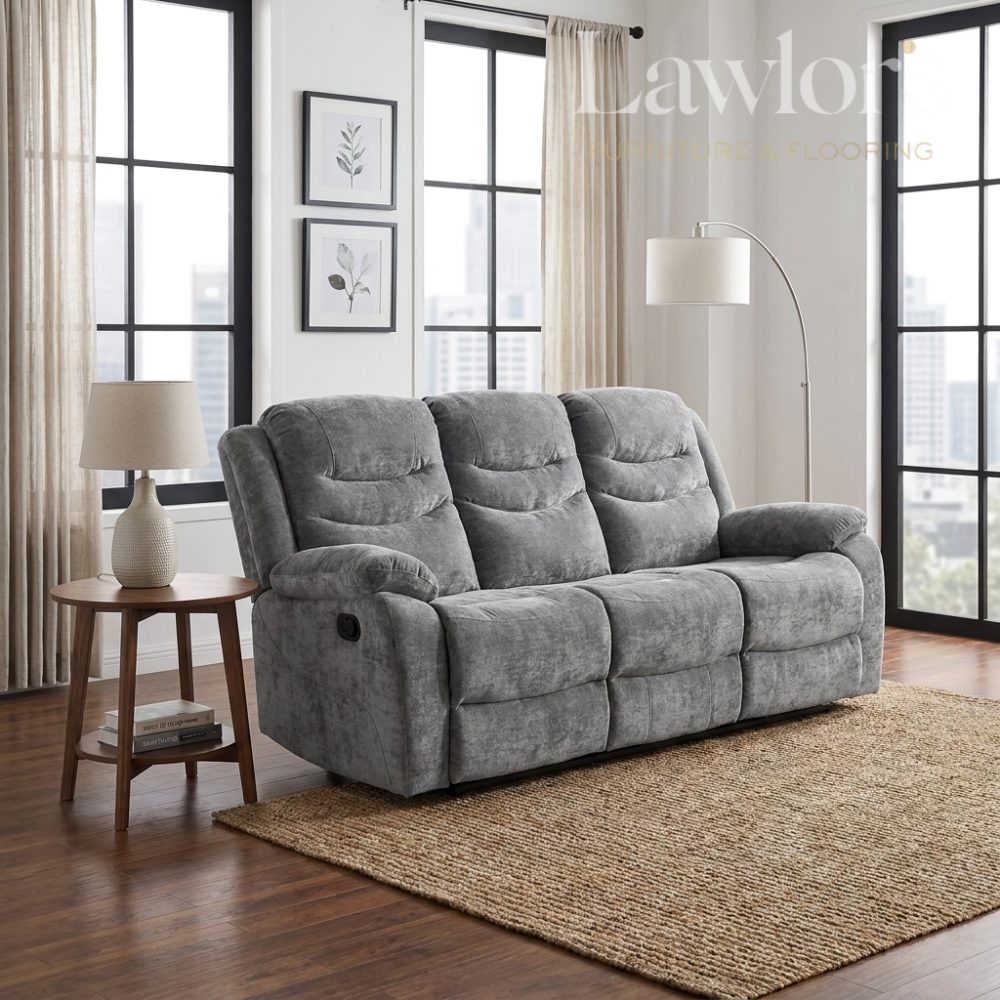 A gray reclining three seater sofa in a modern living room setting with wood floors, a woven rug, a side table with a lamp, and large windows.