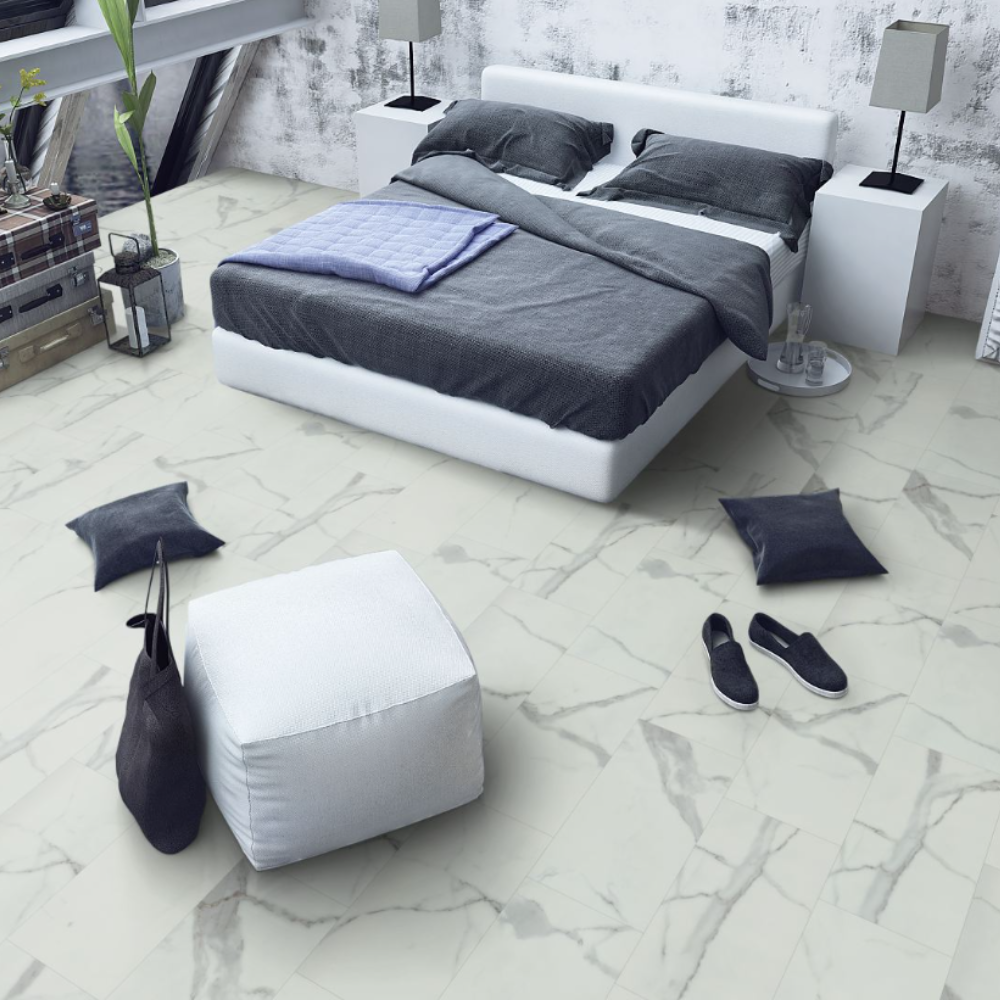 A modern bedroom scene showing a white platform bed with dark gray bedding, a white ottoman, dark throw pillows, and a light marble-look floor. No people or animals are present.