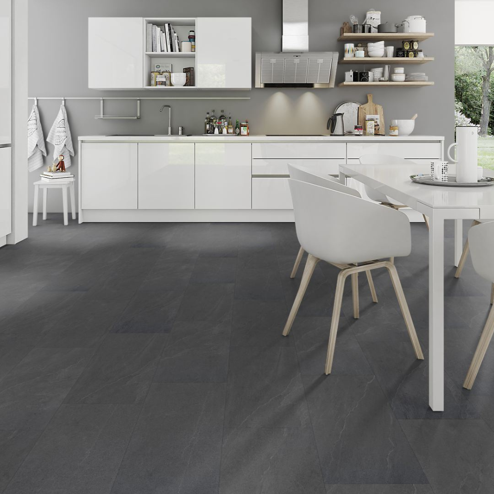 Dark charcoal gray stone-look floor tiles in a modern white kitchen setting, shown from a high angle with white dining chairs and a white table in the foreground.