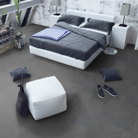 A modern bedroom scene showing a white platform bed with dark gray bedding, a white ottoman, gray floor pillows, bedside tables with lamps, and gray flooring. No people or animals are present.
