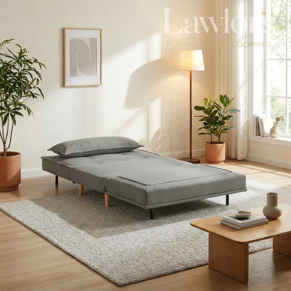 Grey sofa bed in a living room setting with plants and a lamp.