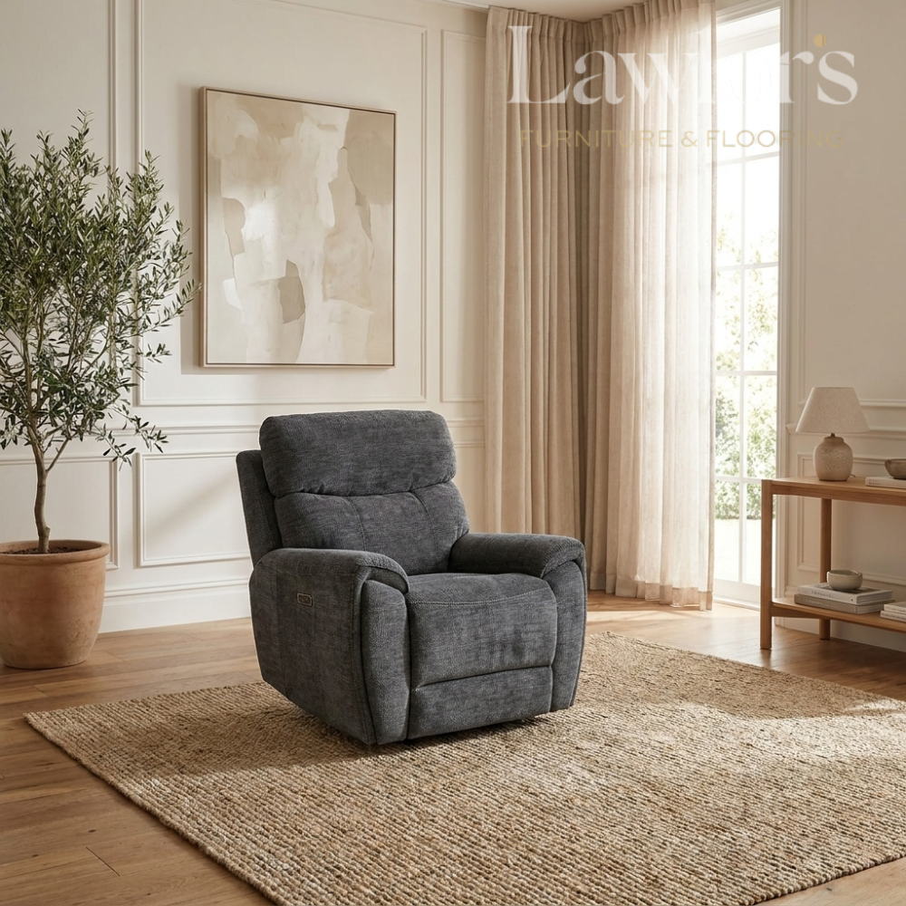A dark gray upholstered recliner chair placed on a woven area rug in a bright, modern living room with wood floors, beige curtains, a potted tree, and wall art.