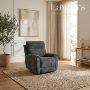 A dark gray upholstered recliner chair placed on a woven area rug in a bright, modern living room with wood floors, beige curtains, a potted tree, and wall art.