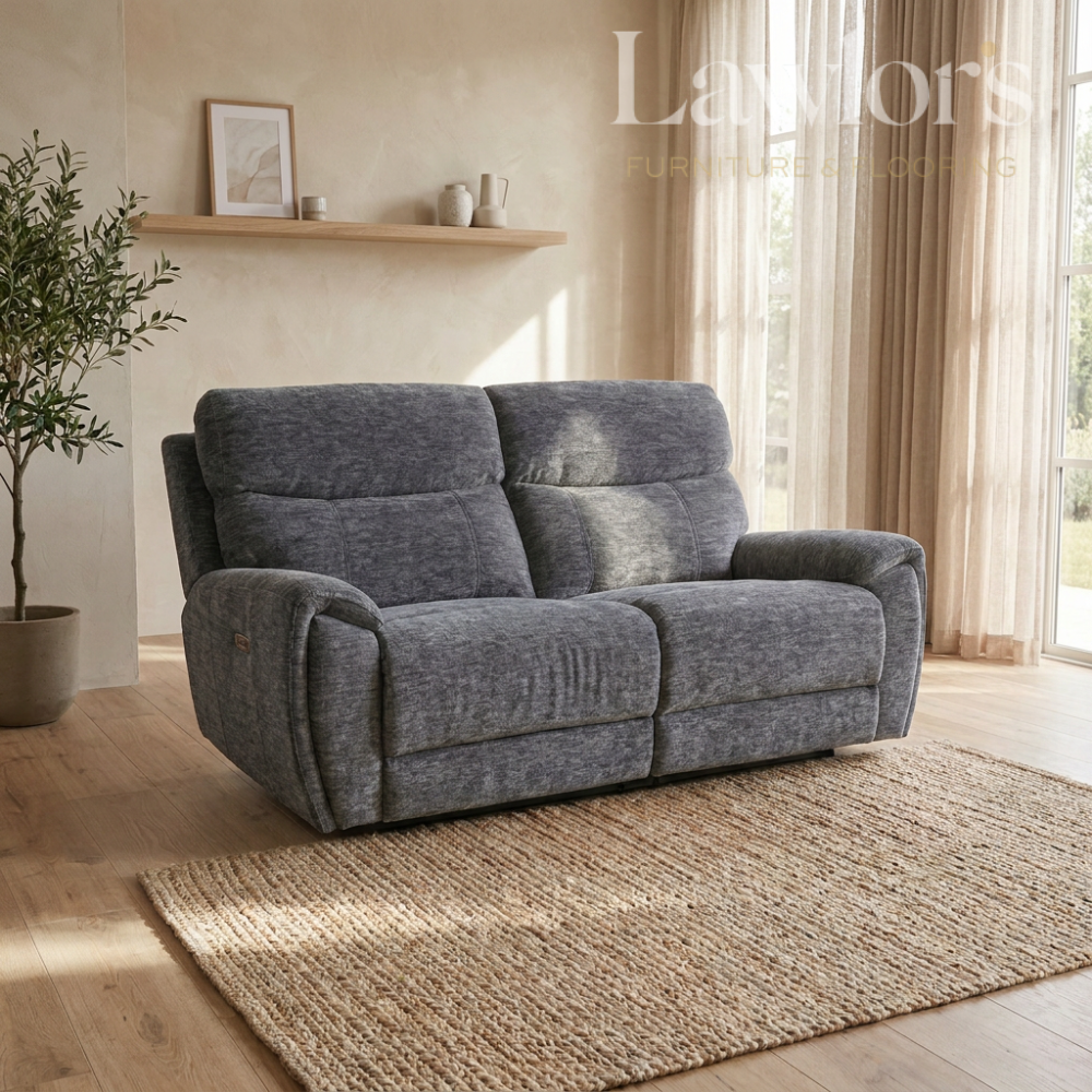 A gray upholstered reclining loveseat sofa placed in a bright living room with wood floors, a woven rug, curtains, and a shelf in the background.