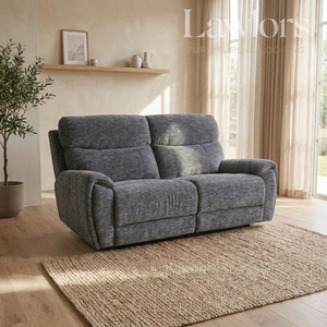 A gray upholstered reclining loveseat sofa placed in a bright living room with wood floors, a woven rug, curtains, and a shelf in the background.