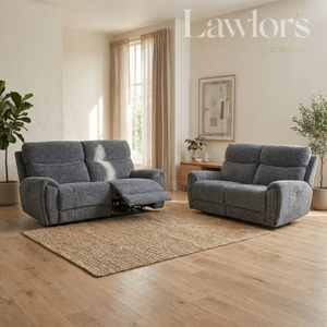 A gray upholstered reclining loveseat and matching armchair shown in a bright living room with wood floors, a woven rug, curtains, and plants.