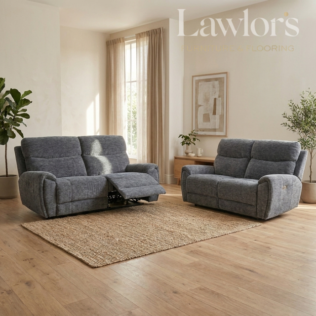 A gray upholstered reclining loveseat and matching armchair shown in a bright living room with wood floors, a woven rug, curtains, and plants.