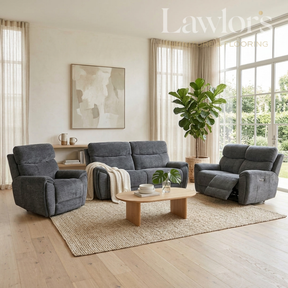 A modern living room set featuring a dark gray three-seater sofa, a matching recliner, and a gray armchair arranged on a woven rug around a light wood coffee table. The room has light wood floors, beige curtains, and large windows with a green plant in the corner.