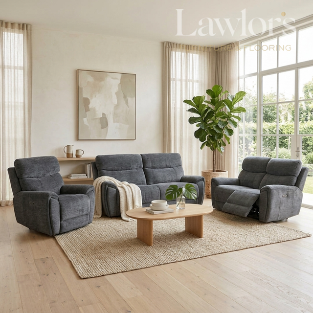 A modern living room set featuring a dark gray three-seater sofa, a matching recliner, and a gray armchair arranged on a woven rug around a light wood coffee table. The room has light wood floors, beige curtains, and large windows with a green plant in the corner.