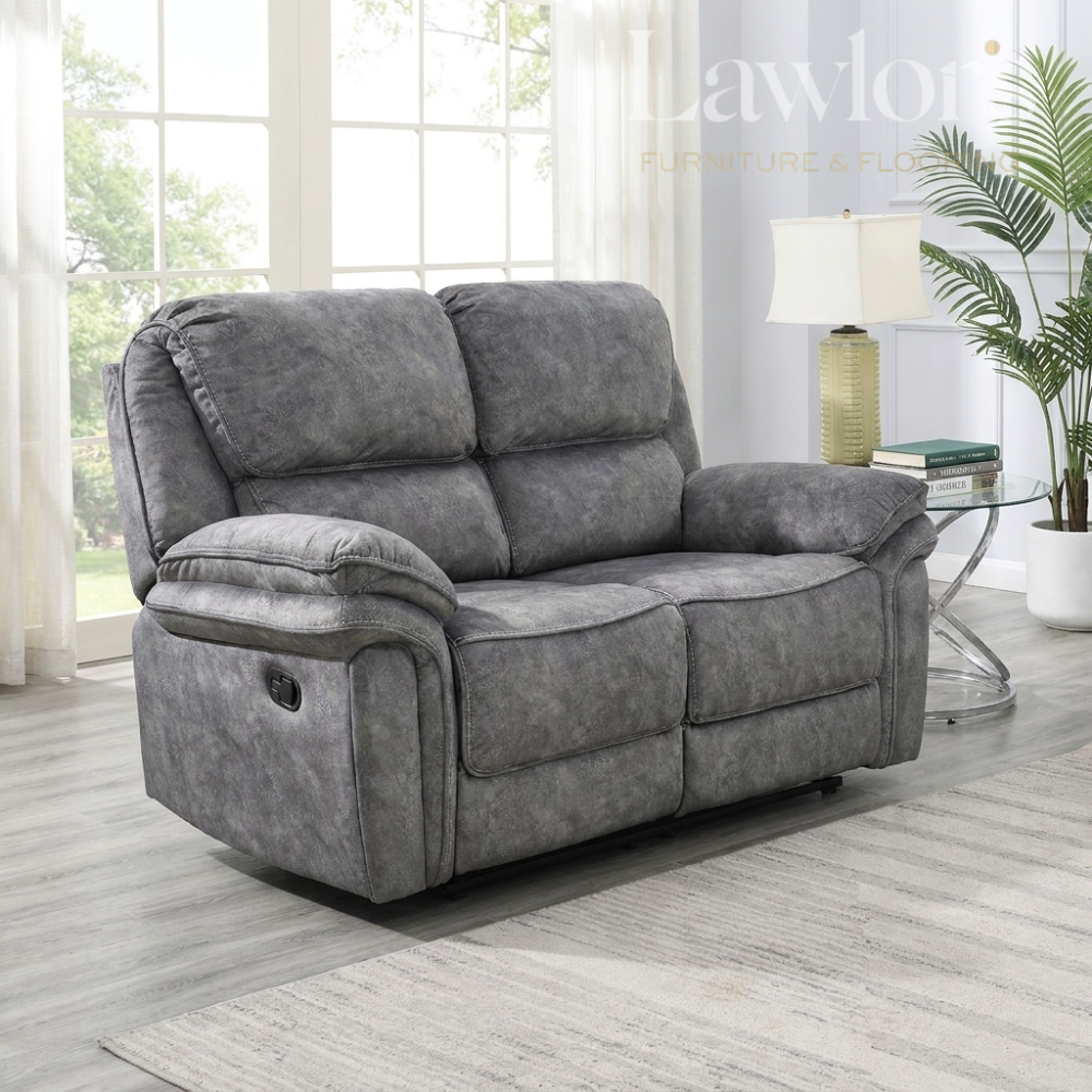 A dark grey upholstered reclining 2 seater shown in a bright living room setting with windows, a side table, lamp, and plant.