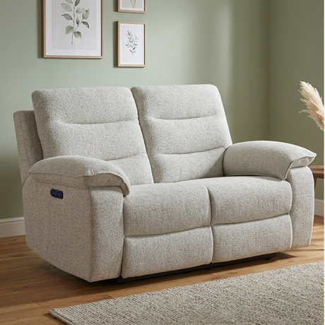 A light beige upholstered reclining loveseat shown in a styled living room with a sage green wall, wood floor, framed botanical prints, and a rug.