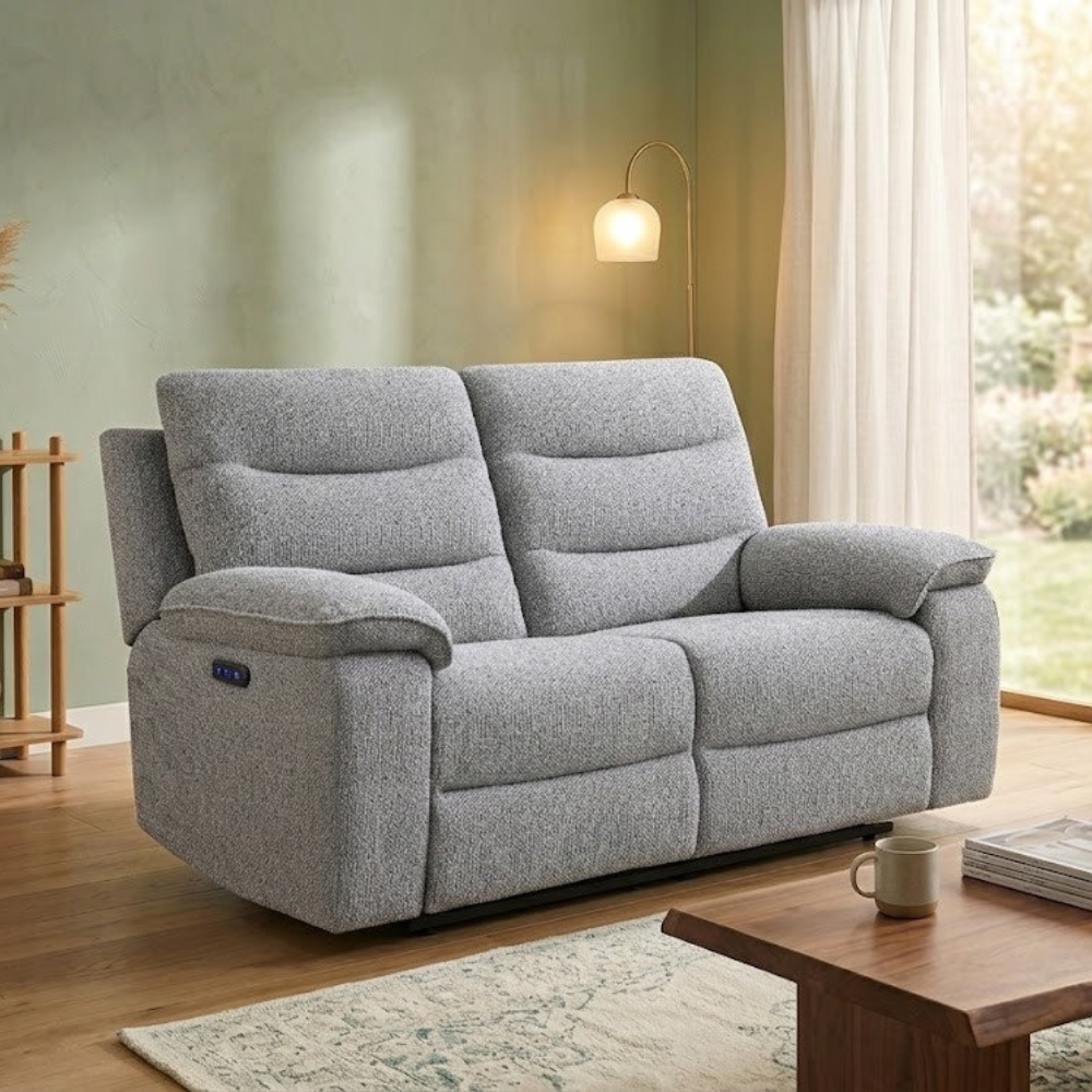 A light gray upholstered reclining loveseat sofa shown in a styled living room with wood flooring, a floor lamp, curtains, and a coffee table.
