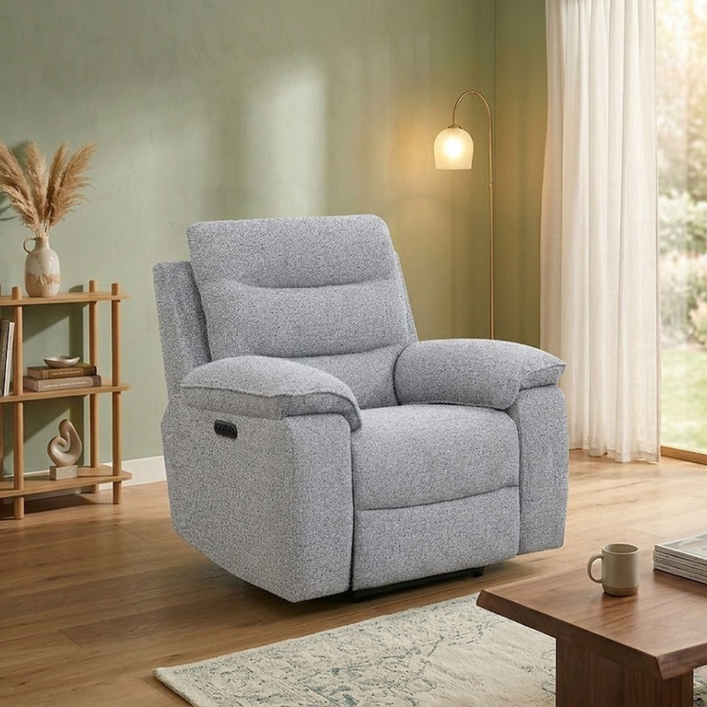 A light gray upholstered recliner chair in a styled living room with wood floors, a green wall, a floor lamp, a small wooden shelf, and a coffee table.