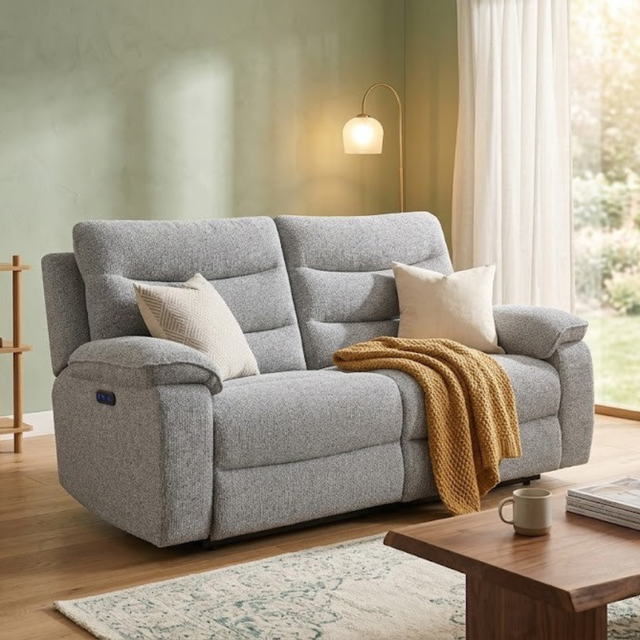 A light gray upholstered reclining loveseat in a styled living room, with two cream throw pillows, a mustard yellow knit blanket draped over the seat, a floor lamp, window, and wooden coffee table nearby.