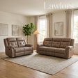 A staged living room scene showing two tufted reclining sofas in a warm taupe-brown upholstery, placed on a large textured rug over light wood flooring. The room has cream walls, beige curtains, a floor lamp, a potted plant, and framed wall art.