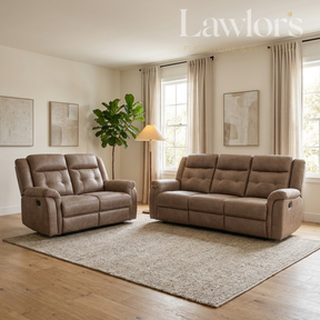 A staged living room scene showing two tufted reclining sofas in a warm taupe-brown upholstery, placed on a large textured rug over light wood flooring. The room has cream walls, beige curtains, a floor lamp, a potted plant, and framed wall art.