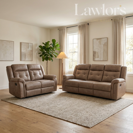 A staged living room scene showing two tufted reclining sofas in a warm taupe-brown upholstery, placed on a large textured rug over light wood flooring. The room has cream walls, beige curtains, a floor lamp, a potted plant, and framed wall art.