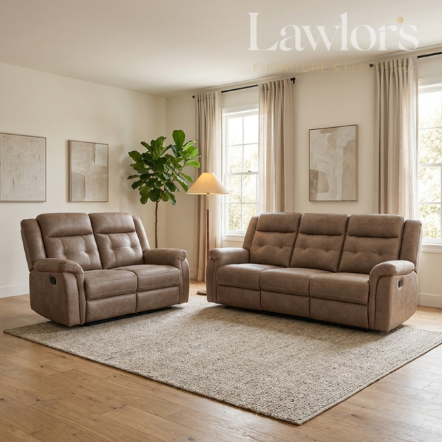 A staged living room scene showing two tufted reclining sofas in a warm taupe-brown upholstery, placed on a large textured rug over light wood flooring. The room has cream walls, beige curtains, a floor lamp, a potted plant, and framed wall art.