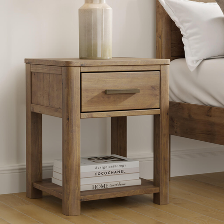Wooden nightstand with a drawer and open shelf, books on a bed in the background