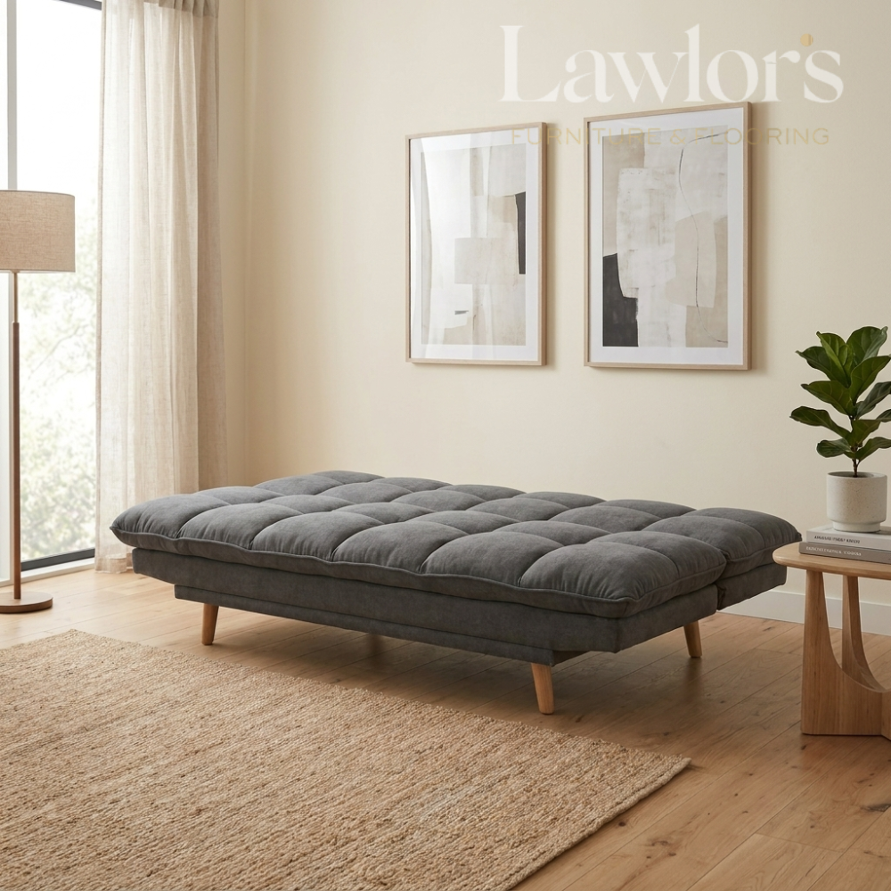 A gray tufted cushioned daybed with light wood legs in a modern, sunlit living room setting. The room features beige walls, a woven rug, a floor lamp, and framed abstract art.