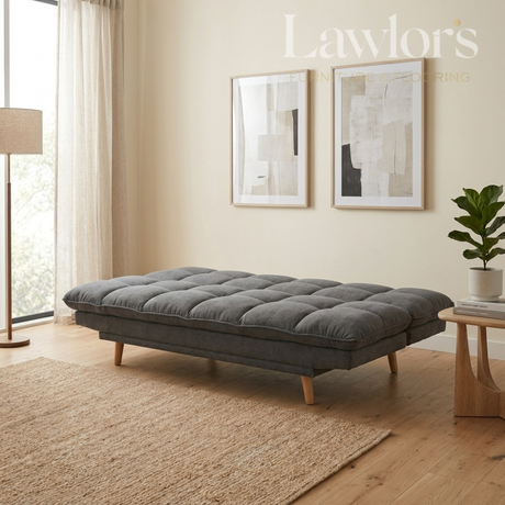 A gray tufted cushioned daybed with light wood legs in a modern, sunlit living room setting. The room features beige walls, a woven rug, a floor lamp, and framed abstract art.