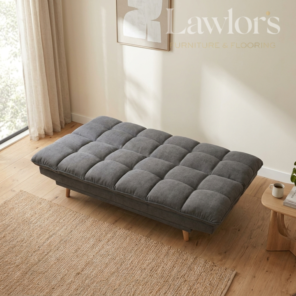 A gray tufted futon sofa bed with light wood legs, styled in a bright, modern room with a beige rug, wooden floor, and a small wooden side table.