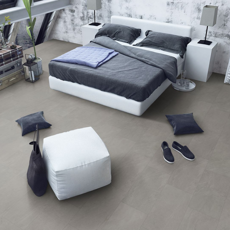 A modern bedroom scene showing a white platform bed with dark gray bedding, flanked by white bedside tables and lamps, with a white ottoman and dark throw pillows on a light gray spc floor.