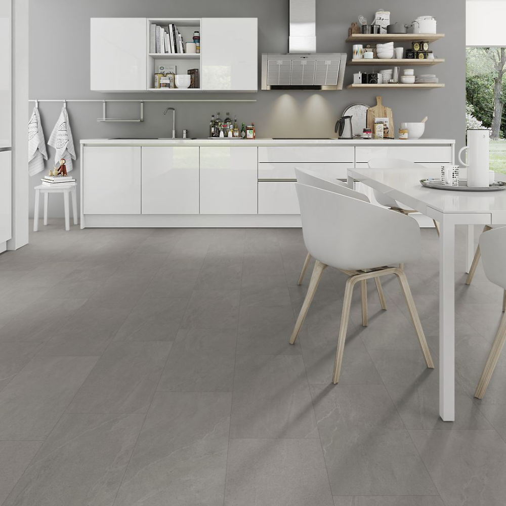 A modern kitchen interior with white cabinets, a white dining table, white chairs with light wood legs, and large gray floor tiles. The image appears to emphasize the spc flooring, with the floor occupying most of the frame and the kitchen serving as contextual staging.