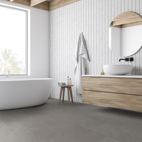 A modern bathroom interior with a freestanding white bathtub, a floating wood vanity with a white vessel sink, white wall tiles, a round mirror, and grey spc flooring. A white towel hangs on a small wooden stool beside the vanity.