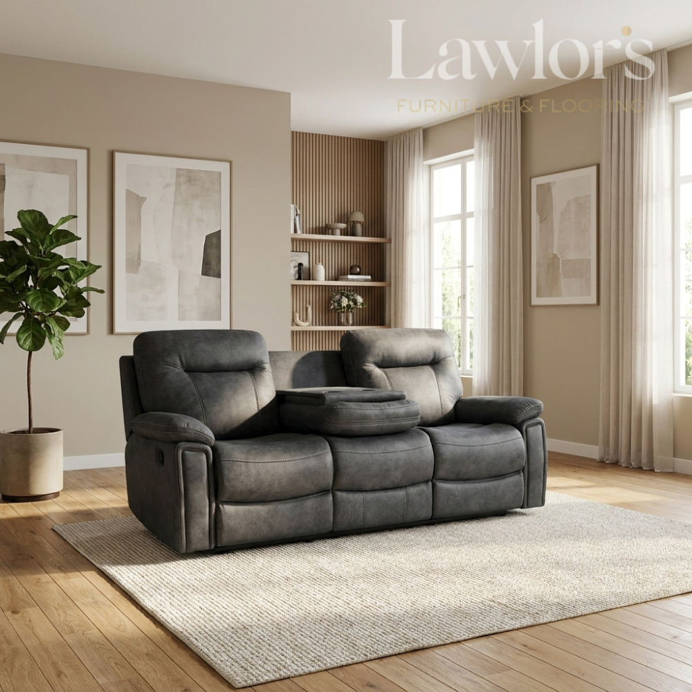 A dark gray two-seater reclining sofa with a central cup holder, styled in a modern living room setting on a light beige rug over a wooden floor. The room features beige walls, framed abstract art, a potted plant, and tall windows with sheer curtains.
