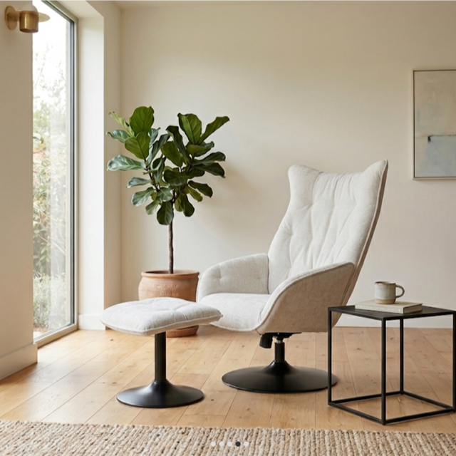 A modern upholstered lounge chair with a matching ottoman in a bright, minimalist room with light wood floors, a potted fiddle-leaf fig, and a small black side table.