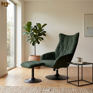 A dark green velvet wingback armchair with a matching ottoman in a bright, modern room with light wood floors, a potted fiddle-leaf fig, and a small side table.