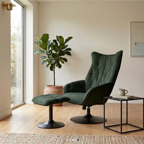 A dark green velvet wingback armchair with a matching ottoman in a bright, modern room with light wood floors, a potted fiddle-leaf fig, and a small side table.