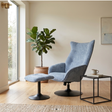 A modern light blue upholstered wingback armchair with a matching ottoman in a bright, minimalist room with a potted fiddle-leaf fig plant and a small black side table.