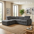 A modern L-shaped sectional sofa in dark gray fabric, styled in a bright, contemporary living room with beige walls, a woven rug, and framed wall art.