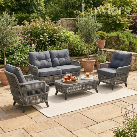Outdoor garden furniture patio set with gray cushions on a stone patio surrounded by greenery.
