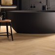 A modern freestanding black bathtub in a minimalist bathroom setting with light wood herringbone flooring, a small wooden stool with a black bottle, and dark wall fixtures.