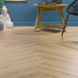 Light oak herringbone wood flooring shown in a styled room setting with a blue wall, a bamboo side table, a white Buddha statue, and a black chair.