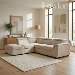 A light beige fabric sectional sofa with a chaise ottoman in a bright, modern living room. The room features light wood floors, a textured cream rug, a wooden sideboard with a potted plant, and large windows with warm natural light.