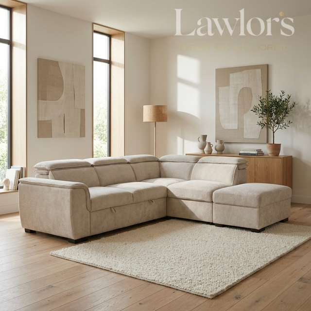 A light beige fabric sectional sofa with a matching ottoman in a bright, modern living room. The room features light wood floors, a textured cream rug, a wooden sideboard with a lamp and plant, and abstract wall art.